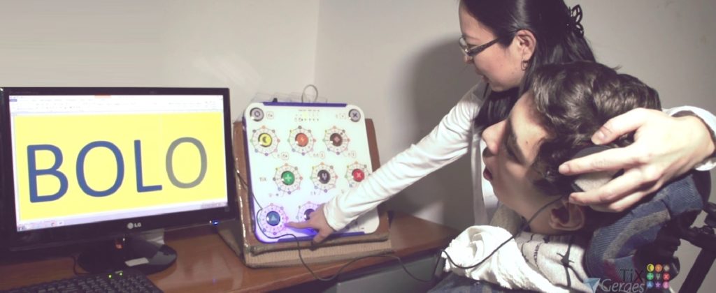 Terapeuta indica uma tecla do TiX para o paciente na cadeira de rodas acionar com a cabeça, fazendo aparecer a palavra BOLO na tela
