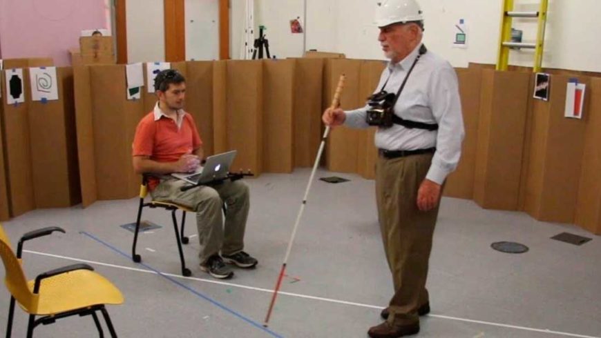 Homem com bengala, capacete e sensor preso ao tórax por tiras caminha em uma sala de piso cinza onde há um homem ao fundo sentado com um notebook no colo