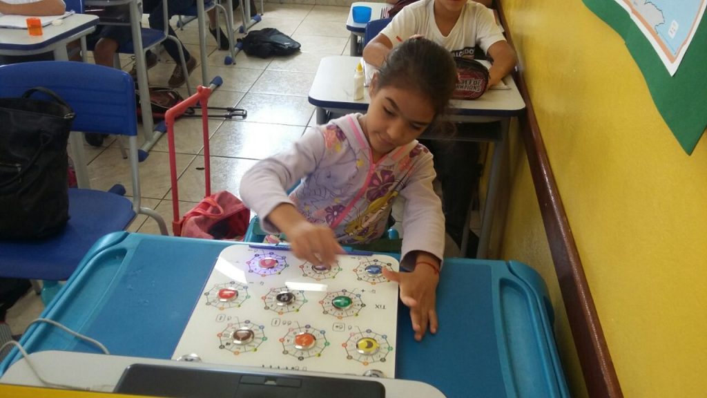 Menina criança tocando as teclas do TiX diante de um notebook sobre uma mesa adaptada, dentro de uma sala de aula regular