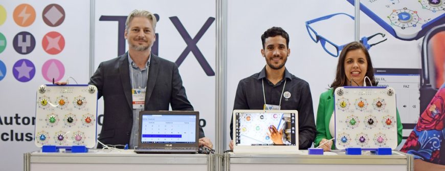 Três pessoas posam no estande da TiX Tecnologia, atrás de balcões onde se encontram os produtos da empresa e notebooks. Da esquerda para direita, posam sorridentes: Marcelo Rubinger, Adriano Assis e Ana Nasquewitz.