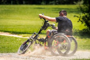 homem pilotando uma cadeira de rodas adaptada como um triciclo elétrico sobre uma pista de terra com um gramado ao fundo