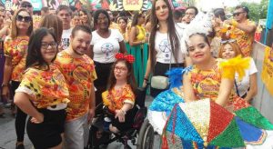 Varias pessoas do bloco de carnaval posando para a foto, entre elas pessoas com síndrome de down e uma cadeirante. Todos sorrindo e usando roupas carnavalescas