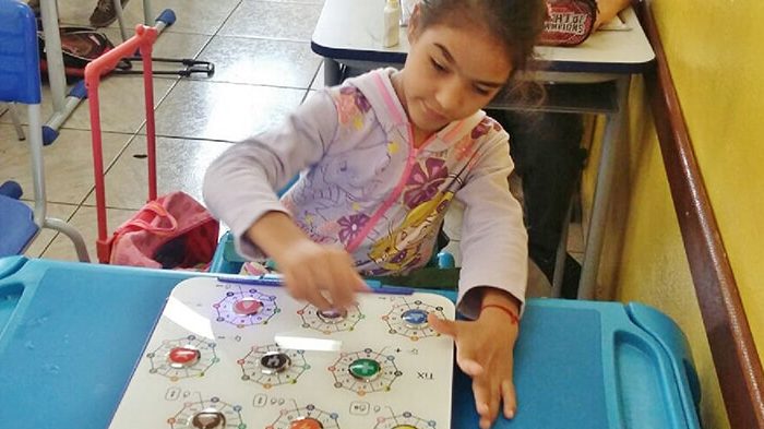 Menina Julia, que tem paralisia cerebral, utilizando o Teclado TiX sobre uma mesa escolar em uma sala de aula.