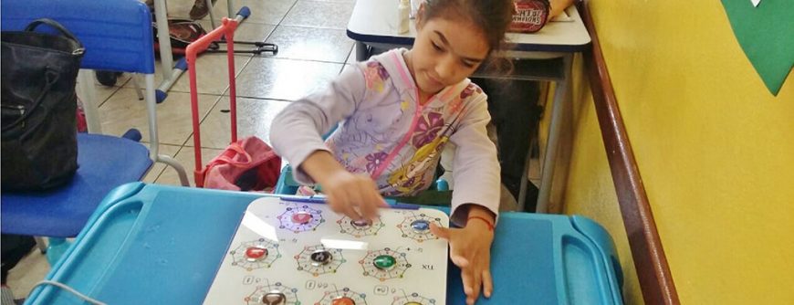 Menina Julia, que tem paralisia cerebral, utilizando o Teclado TiX sobre uma mesa escolar em uma sala de aula.