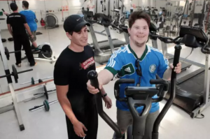 Rapaz com síndrome de down se exercitando no transport em uma academia. Com o professor em pé ao seu lado.